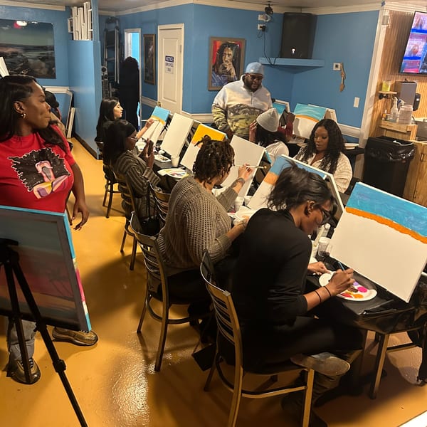 People painting on canvases at tables in a blue-walled room with a Bob Marley portrait.