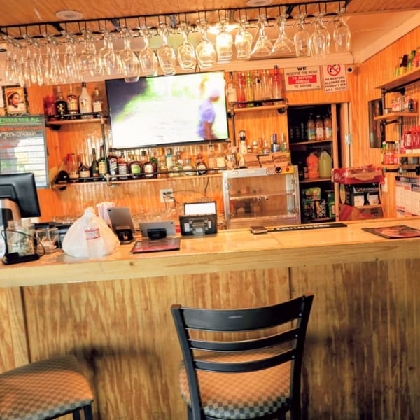 A Jamaican restaurant bar with liquor bottles, hanging glasses, a TV, and two bar stools.