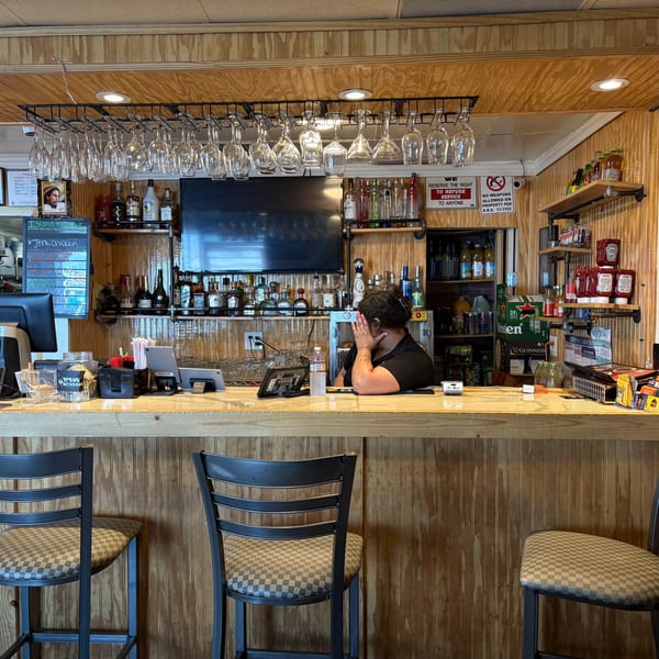 A bartender rests her head on her hand behind a wooden bar with bottles and hanging glasses.