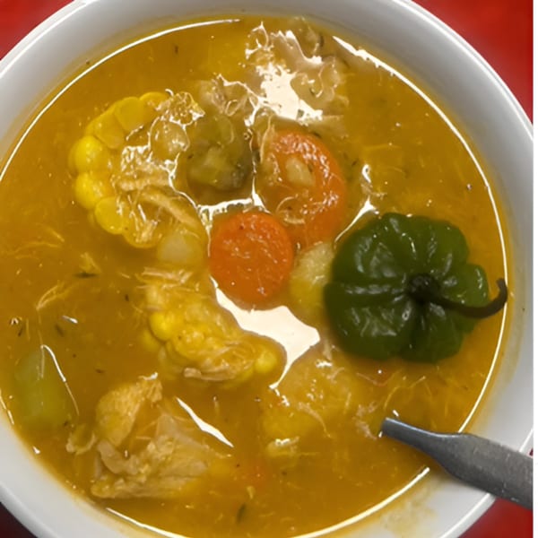 A bowl of Jamaican soup with corn, carrots, and a green Scotch bonnet pepper.